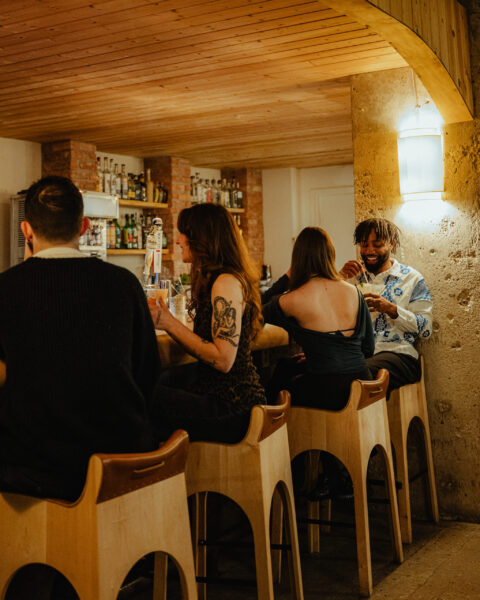 People sitting on wooden stools chat and enjoy drinks at a cozy, dimly lit bar with a wooden ceiling.