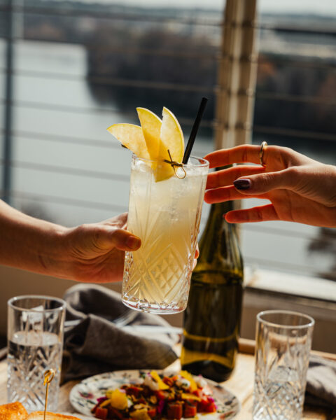 Hands toasting with a citrus cocktail over a table of colorful dishes
