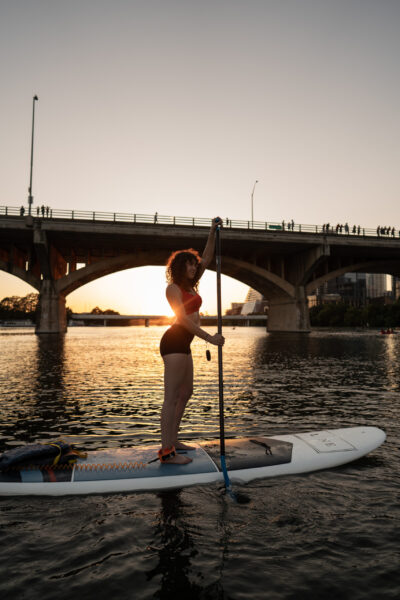 Woman stand-up paddleboarding on a river at sunset, with a large arched bridge in the background.