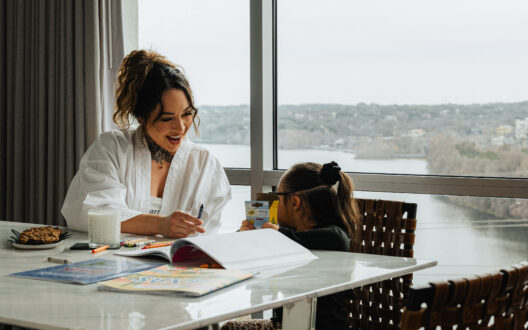 A woman and child color together at a table by a large window overlooking a river valley.