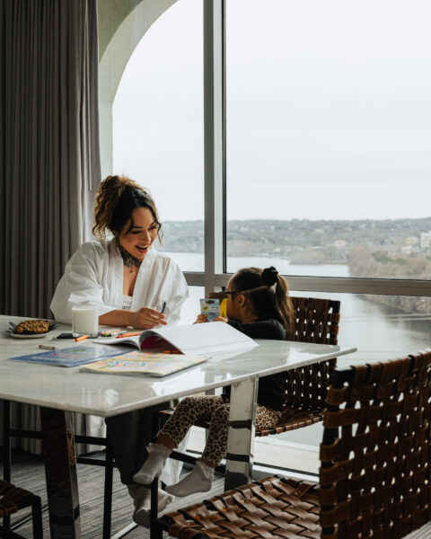 A woman and child color together at a table by a large window overlooking a river valley.