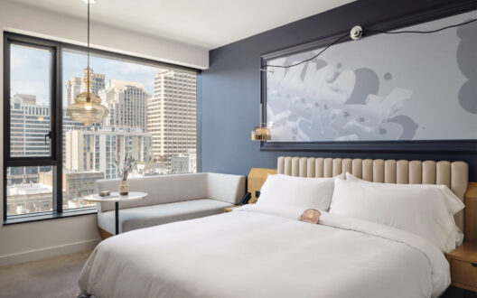In the hotel room, a bed and a couch are placed in the right corner, near the glass window with a wonderful city view.